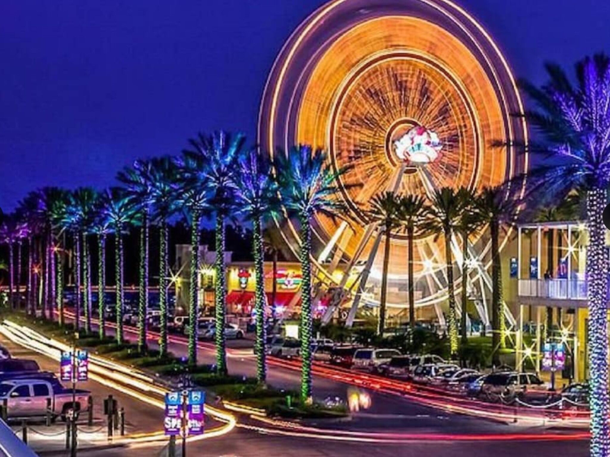 A nighttime view of a large Ferris wheel with palm trees lining the street.