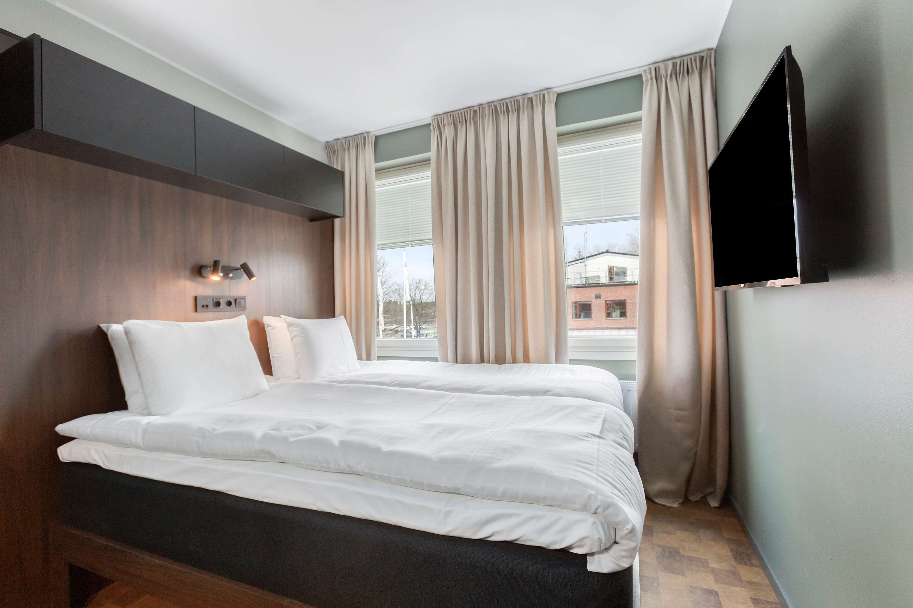 A modern hotel room with a large bed, a flat-screen TV mounted on the wall, and a window with curtains.