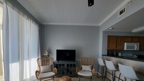 A living room with a TV, two wicker chairs, a coffee table, a striped rug, and a kitchen area with a microwave. - Inn at Dauphin Island 306 Efficient 1BR sleeps 6 with Beautiful Views!! (Dauphin Island)