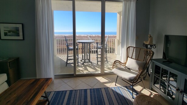 A room with a sliding glass door leading to a balcony with a view of the beach. There is a table and a chair on the balcony, and a television on a cabinet. - Inn at Dauphin Island 306 Efficient 1BR sleeps 6 with Beautiful Views!! (Dauphin Island)