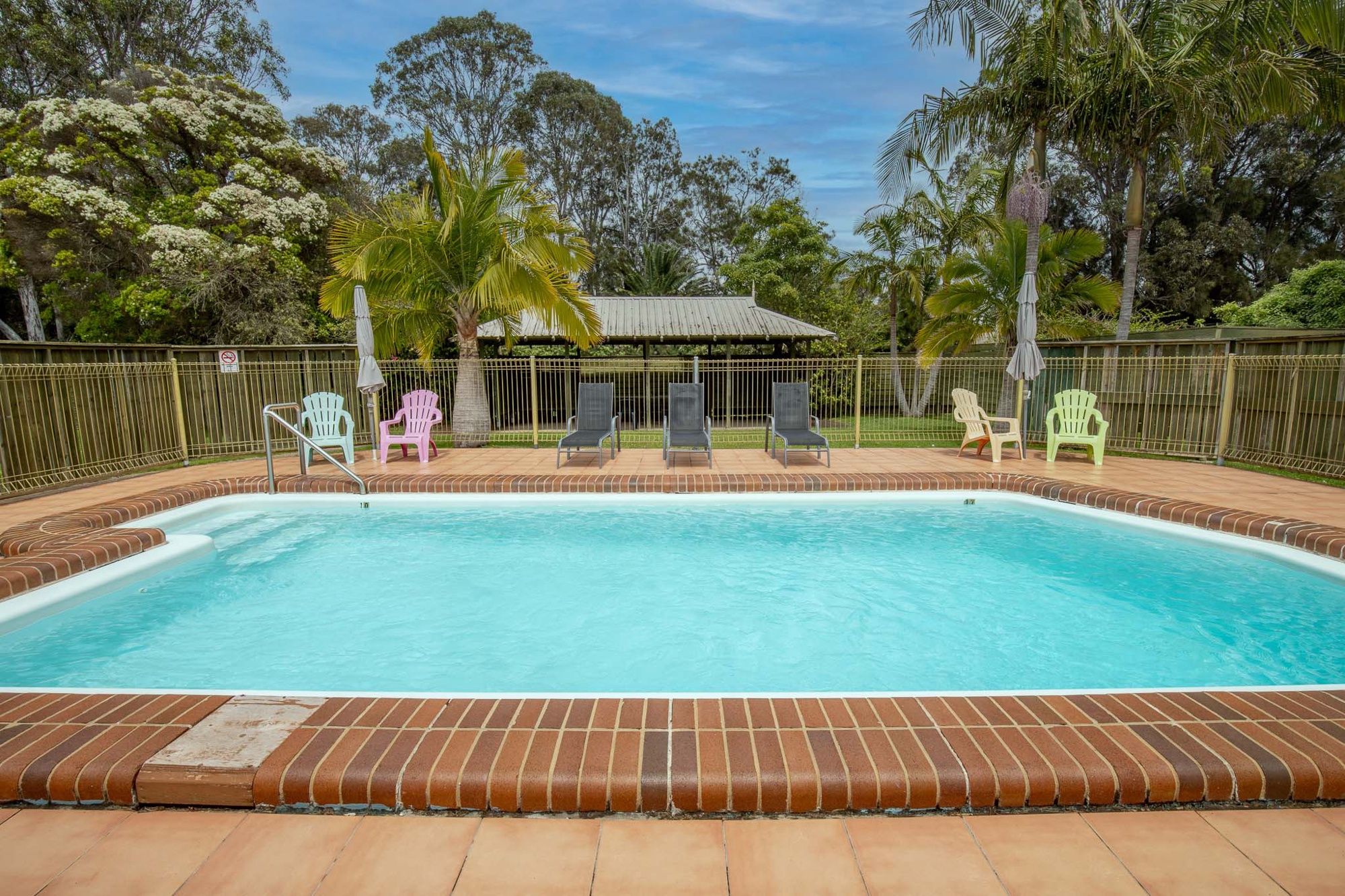 Une piscine rectangulaire bordée de briques, entourée d’une clôture, et des chaises sont disposées autour de la piscine.