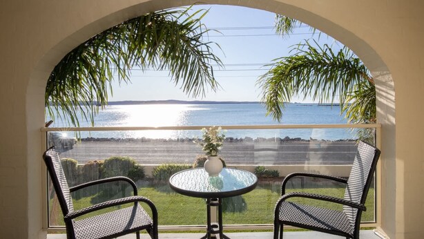 A balcony with two wicker chairs and a glass table, overlooking a body of water and a distant shoreline. - Seafront Getaway Studio, Great Location with Pool (Batemans Bay)
