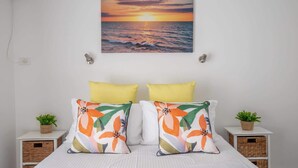 A bedroom with a bed, two pillows, a wall-mounted picture of a sunset over the ocean, and two nightstands with small potted plants. - Seafront Getaway Studio, Great Location with Pool (Batemans Bay)