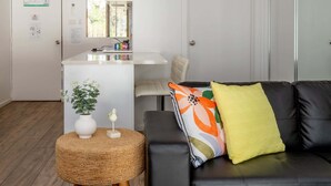 A modern living room with a black leather sofa, a woven ottoman, and a small white table with a plant. A kitchen area is visible in the background with a window and a refrigerator. - Seafront Getaway Studio, Great Location with Pool (Batemans Bay)