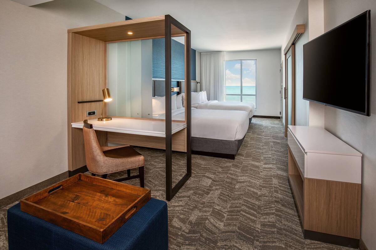 Habitación de hotel con escritorio, silla y cama. Hay un televisor montado en la pared y una ventana con vistas al océano.