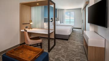 Quarto de hotel com escrivaninha, cadeira e cama. Há uma televisão na parede e uma janela com vista para o oceano.