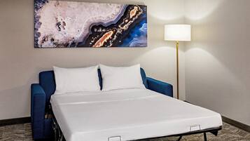 A hotel room with a bed, a blue armchair, a white lamp, and a large abstract painting on the wall.