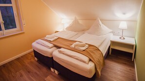 Una cama bien tendida con almohadas y una manta, suelo de madera, una ventana y lámparas de noche.