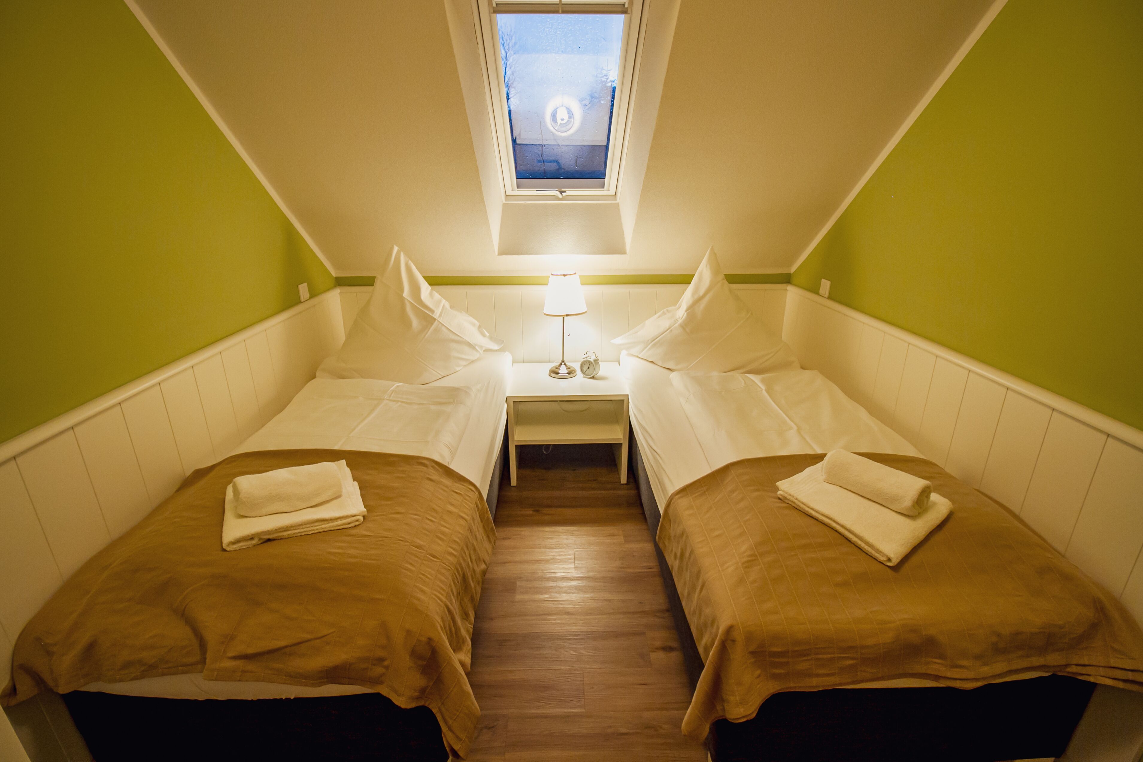 Une petite chambre d’hôtel composée d’une seule pièce, avec deux lits, une table de chevet, une lampe et une fenêtre donnant sur la rue.