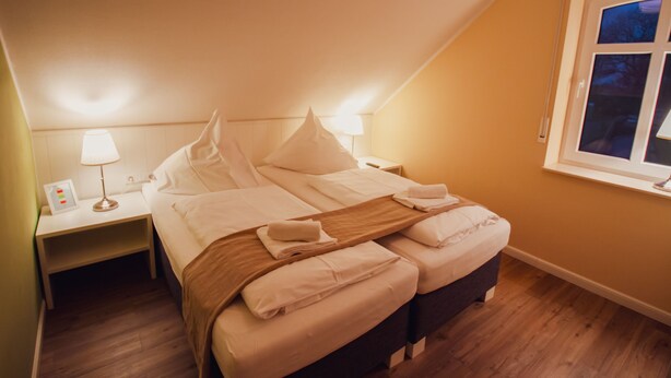 Ein ordentlich bezogenes Bett in einem Zimmer mit Holzboden, einem Nachttisch mit Lampe und einem Fenster mit Ausblick.