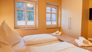 A hotel room with two beds, a window with blinds, a wooden bench, and a towel on the bench. - Int Corner 6 (Krummhörn)