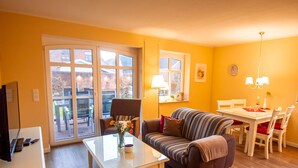 A living room with a sofa, a coffee table, and a dining table with chairs. There is a TV on a stand, a window with curtains, and a lamp on the dining table. - Int Corner 6 (Krummhörn)