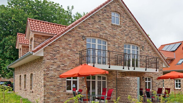 A two-story brick house with a balcony, outdoor seating with red chairs and umbrellas, and a visible garage. - Int Corner 6 (Krummhörn)