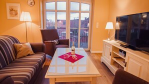 A living room with a sofa, armchair, glass coffee table, TV, and a balcony with a view. - Int Corner 7 (Krummhörn)