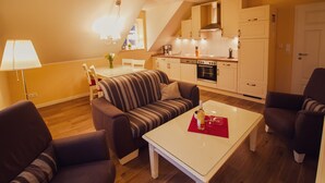 Una sala moderna con sofá, sillones y una mesa de centro. Se ve una cocina al fondo con armarios y electrodomésticos blancos.