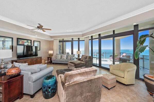 A spacious living room with a large flat-screen TV, comfortable seating, and a balcony with a view of the ocean.