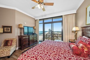 A hotel room with a large bed, a television, a desk with a computer, and a view of the beach.