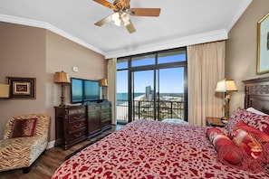 A hotel room with a large bed, a television, a desk with a computer, and a view of the beach.