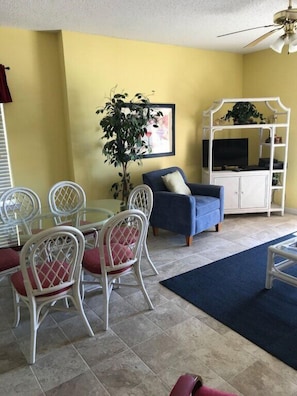 Una sala de estar con pared amarilla, sofá azul, mueble blanco con televisor y comedor con sillas y mesa blancos.