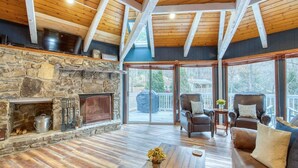 A spacious living area with a stone fireplace, wooden beams, and large windows offering a view of the outdoors. - Balsam Retreat -Charming, Stylish, Great Location! (Maggie Valley)