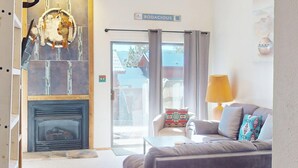 Ein gemütliches Wohnzimmer mit Kamin, einer Couch mit gemusterten Kissen, einer Lampe und Blick ins Freie durch eine Schiebetür.