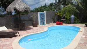 A swimming pool with a curved edge, surrounded by stone tiles, a thatched umbrella, and a potted plant. - Prestige villa with pool and jacuzzi (LE MOULE)