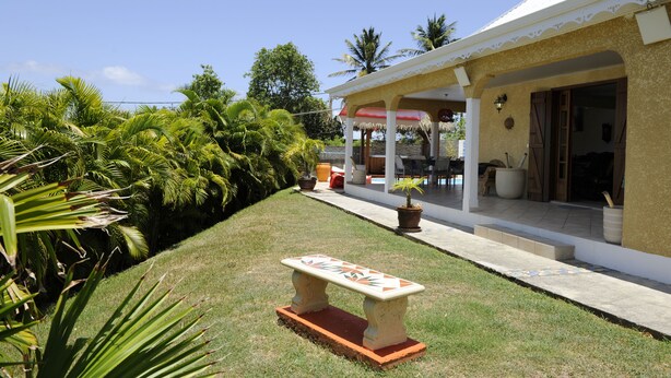 A house with a covered porch, a garden with palm trees, and a stone bench. - Prestige villa with pool and jacuzzi (LE MOULE)