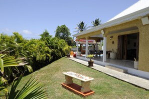 A house with a covered porch, a garden with palm trees, and a stone bench.