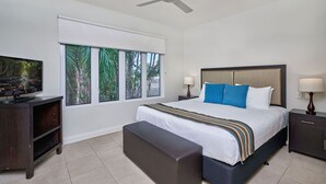 A bedroom with a bed, a TV, a ceiling fan, a window with a view of trees, and a nightstand with a lamp.