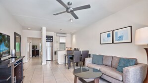 A modern living room with a glass coffee table, a sofa with patterned cushions, a ceiling fan, and a kitchen area in the background.