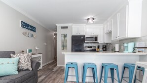 A modern kitchen with white cabinets, a central island with stools, and a grey sofa with patterned cushions. - Newly renovated beautiful Oceanfront Oasis with direct beach access! HEATED POOL (Galveston)