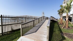 A wooden boardwalk leading to a beach, with a Ferris wheel in the background and a building with a balcony. - Newly renovated beautiful Oceanfront Oasis with direct beach access! HEATED POOL (Galveston)
