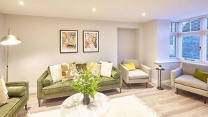 Una sala de estar con un sofá verde, un sillón blanco, una mesa de centro redonda con un jarrón de flores y una lámpara de pie.