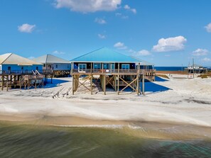Um imóvel à beira-mar com várias estruturas de madeira, incluindo um edifício com telhado azul e varanda, apoiado em estacas sobre a areia.