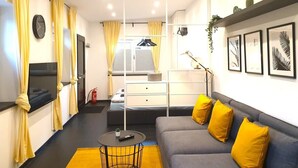 En moderne stue med en grå sofa, gule puder, et sort sofabord og et fladskærms-tv. Der er et vindue med gardiner, en vægmonteret hylde med dekorative genstande og en samling indrammede billeder.