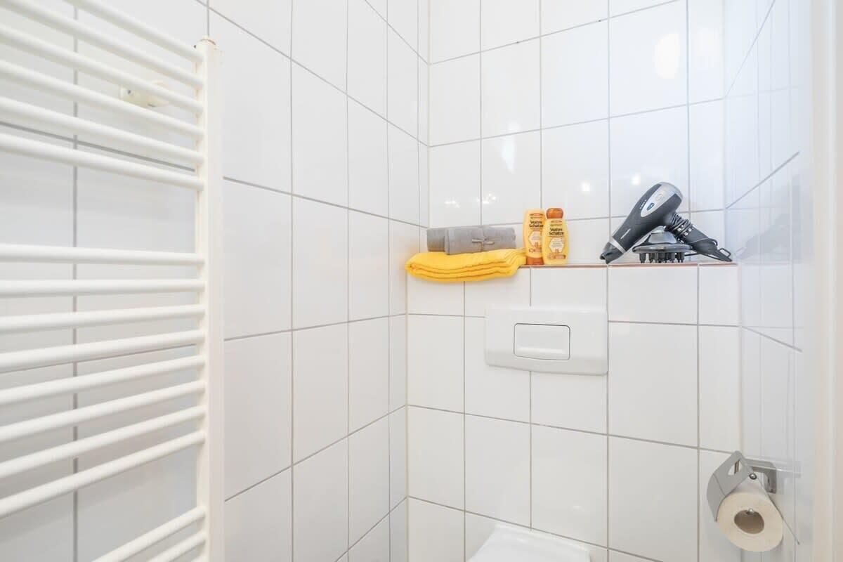 Ein Badezimmer mit weißen Fliesen, einem Handtuchhalter, einem Ablagefach für Toilettenartikel und einem Toilettenpapierhalter.
