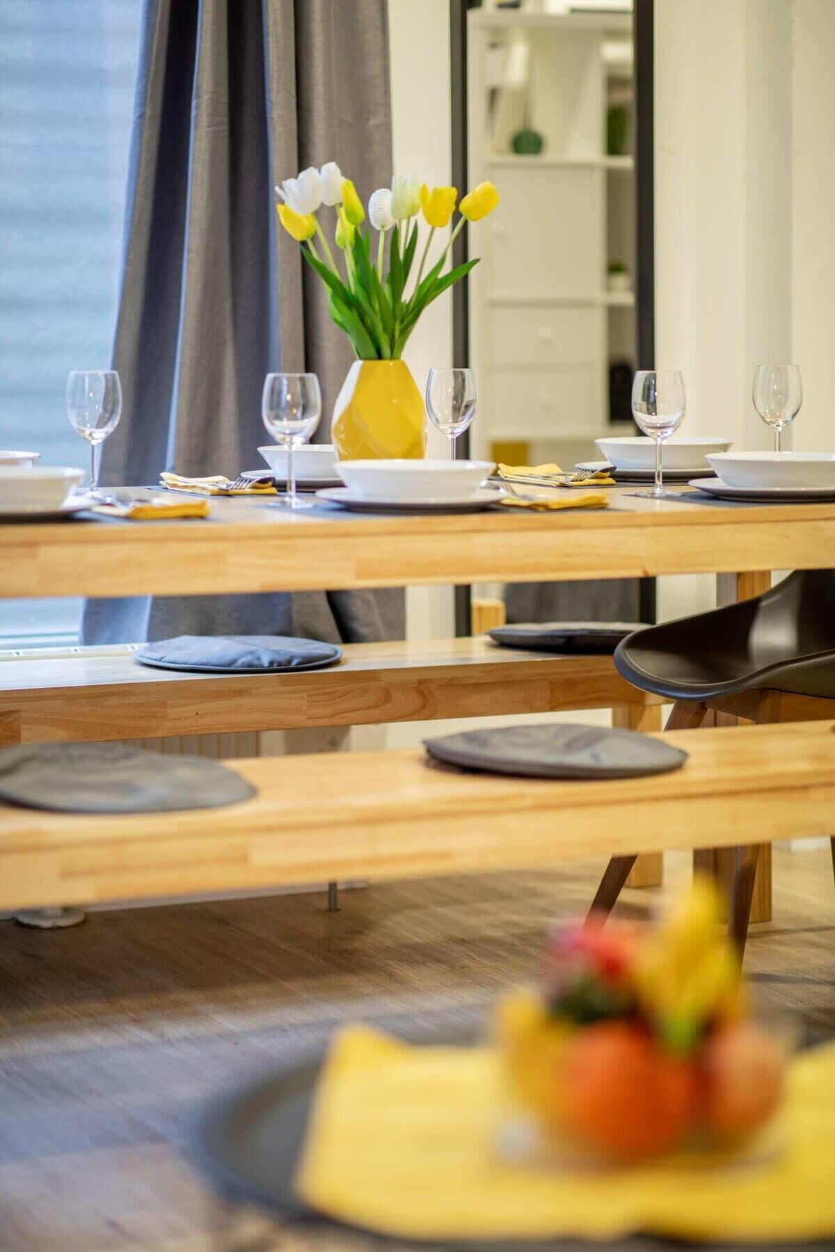 A dining area with a wooden table set for two, a vase with flowers, and a view of the room.