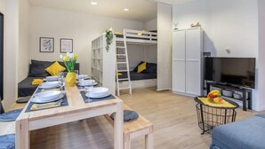 Un espacio habitable compacto con una litera, un comedor con una mesa para dos personas, un sofá, un televisor y una pequeña cocina.