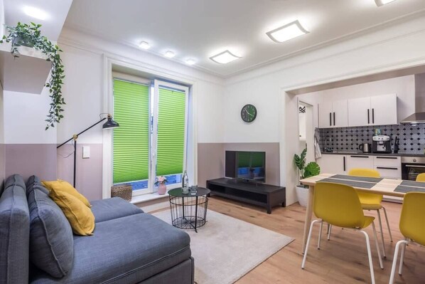 Un salon moderne avec un canapé gris, des coussins jaunes, une petite table ronde et un téléviseur. On y trouve également un coin cuisine avec des meubles blancs et un plan de travail noir.