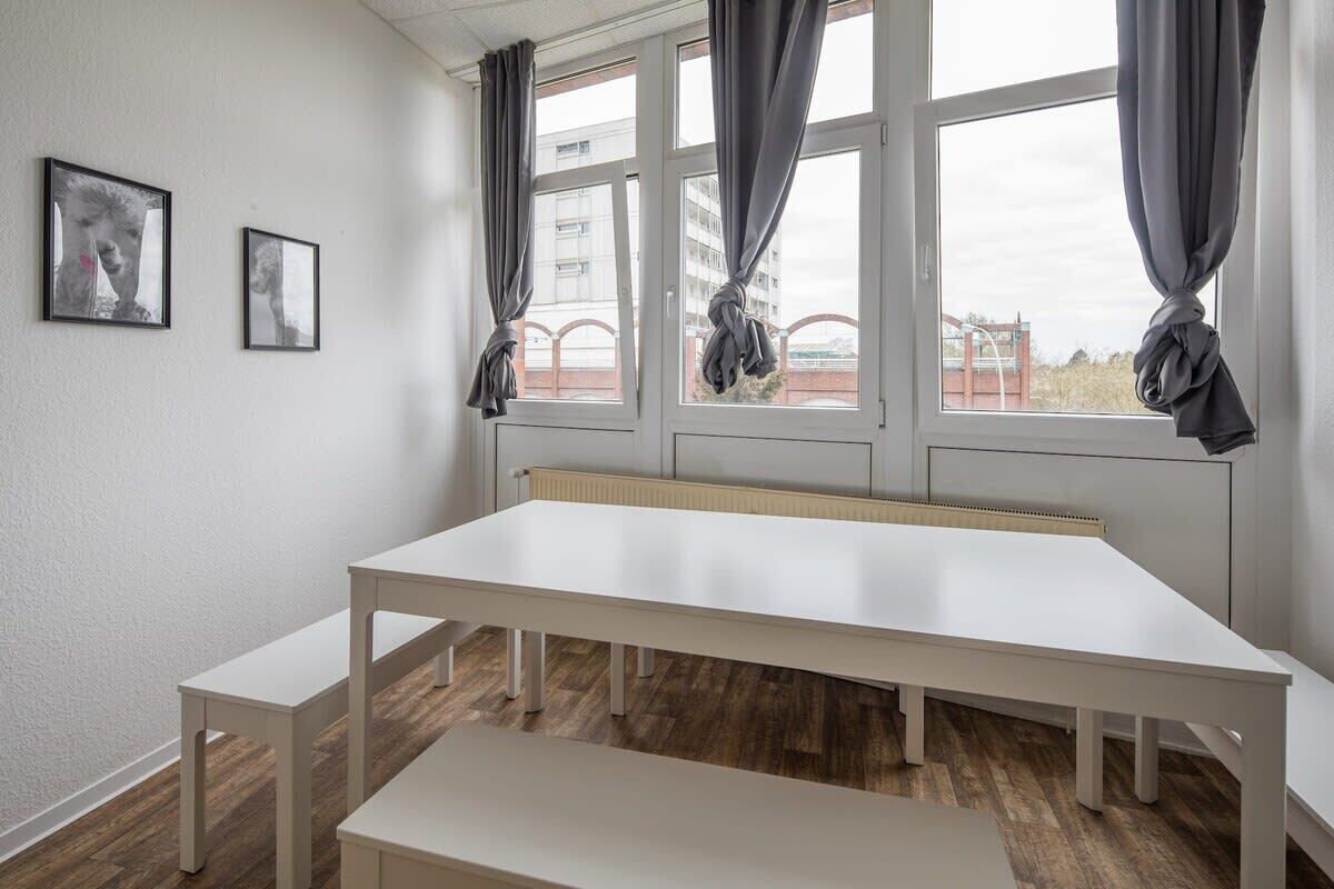 Una habitación con una mesa blanca grande, dos bancos blancos y una ventana con cortinas grises.