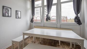 Una habitación con una mesa blanca grande, dos bancos blancos y una ventana con cortinas grises.