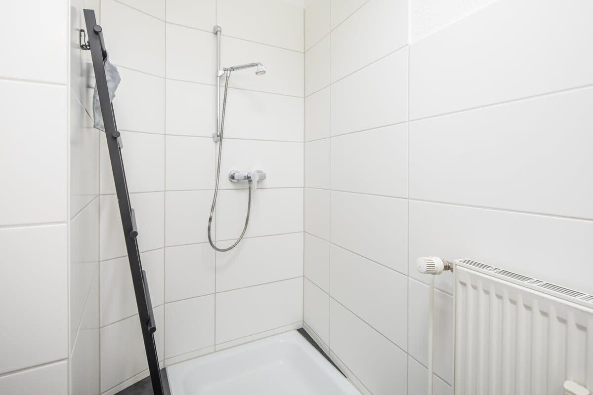 Ett litet duschutrymme med vita kakelplattor, en duschslang och ett vertikalt placerat handtag som man kan hålla sig i.