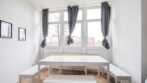 Une pièce avec une grande table blanche, quatre bancs et des rideaux gris aux fenêtres.
