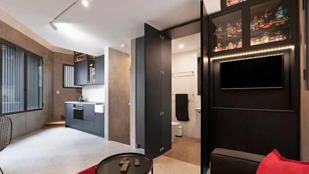 Ein modernes Wohnzimmer mit einem roten Teppich, einem schwarzen Sofa, einem an der dunklen Wand befestigten Fernseher und einer Küche im Hintergrund.