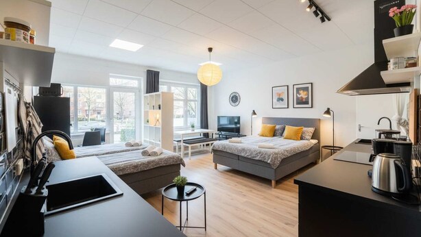 A modern hotel room with a large bed, a desk, a kitchenette, and a view of the outside.