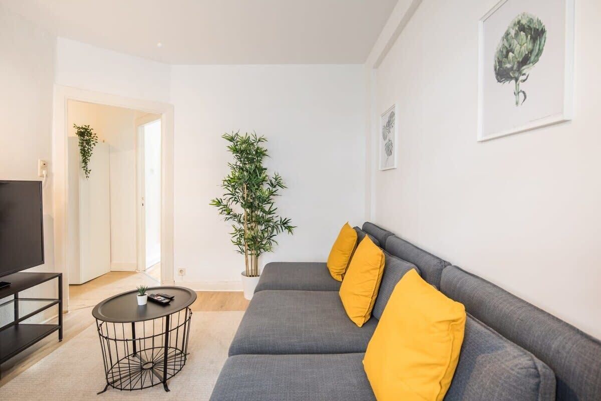 Ein modernes Wohnzimmer mit einem grauen Sofa, gelben Kissen, einem schwarzen Couchtisch, einer Topfpflanze und einem gerahmten Kunstwerk an der Wand.