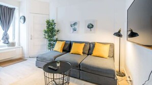 Een moderne woonkamer met een grijze bank, gele kussens, een klein rond tafeltje en een vloerlamp.
