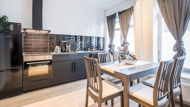 Een moderne keuken met zwarte kasten, een ingebouwde oven, en een eethoek met houten stoelen en een tafel.