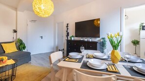 Un salon moderne comprenant un coin repas, un canapé avec des coussins jaunes, une table à manger dressée avec des assiettes et des couverts, et un grand téléviseur à écran plat fixé au mur.