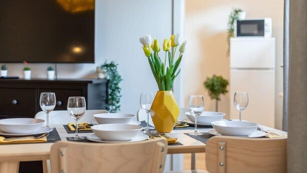 Une table à manger dressée avec de l’assiette blanche, des bols et des couverts, un vase jaune contenant des tulipes, et un téléviseur à écran plat fixé au mur.